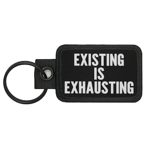 May include: Black rectangular keychain with the text "EXISTING IS EXHAUSTING" in white. The keychain has a black ring and a black fabric loop for attachment.