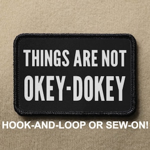 May include: Black patch with white text that reads "THINGS ARE NOT OKEY-DOKEY". The patch has a black border and is designed to be attached with hook-and-loop or sewn on.