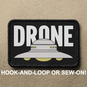 May include: Black patch with white text that reads "DRONE" and a grey UFO with green lights. The patch is for hook-and-loop or sew-on.