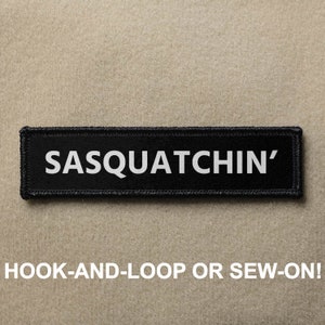 May include: Black patch with white text that reads "SASQUATCHIN'" with hook-and-loop or sew-on backing.