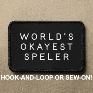 May include: Black patch with white text that reads "WORLD'S OKAYEST SPELER". The patch is for hook-and-loop or sew-on.
