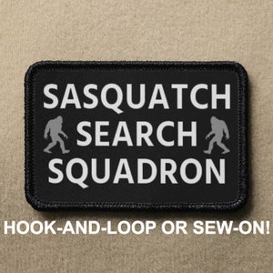 Puede incluir: Parche negro con texto blanco que dice "Sasquatch Search Squadron" con una silueta de Bigfoot a cada lado del texto. El parche tiene un borde negro y está diseñado para ser enganchado o cosido en la ropa.