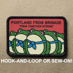 May include: A rectangular patch with a cartoon design of green frogs wearing blue neckties. The patch has a red background with the text "PORTLAND FROG BRIGADE" and "FROG TOGETHER STRONG". The bottom of the patch reads "HOOK-AND-LOOP OR SEW-ON!"