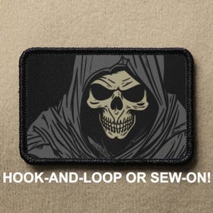 May include: Black and gray embroidered patch featuring a hooded skull with a menacing expression. The patch has a black border and is designed to be sewn or attached with hook-and-loop.