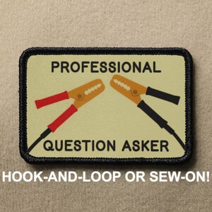 May include: A beige patch with black stitching and the text "PROFESSIONAL QUESTION ASKER" in black. Two red and black jumper cables are shown crossing in the center of the patch. The text "HOOK-AND-LOOP OR SEW-ON!" is printed below the patch.