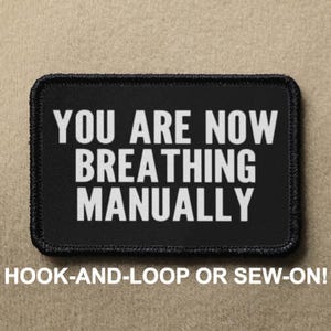 May include: Black rectangular patch with white text that reads "YOU ARE NOW BREATHING MANUALLY." The bottom of the patch says "HOOK-AND-LOOP OR SEW-ON!" The patch has a black border and is on a tan background.