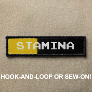 May include: Black and yellow patch with white text that reads "STAMINA" in a pixelated font. The patch is designed to be attached with hook-and-loop or sewn on.