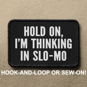 May include: Black patch with white text that reads "HOLD ON, I'M THINKING IN SLO-MO". The patch is for hook-and-loop or sew-on attachment.