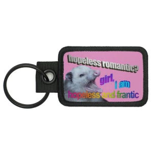 Op de afbeelding: Een roze en zwarte sleutelhanger met een wit opossum met zijn mond open. Het opossum bevindt zich op een roze achtergrond met de tekst "hopeless romantic? girl, i am hopeless and frantic".