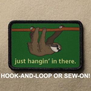 May include: A green and brown embroidered patch with a sloth hanging upside down from a branch. The text "just hangin' in there." is below the sloth. The patch is for hook-and-loop or sew-on attachment.