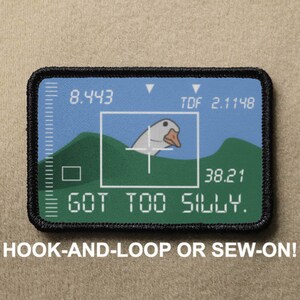 May include: A black and green embroidered patch featuring a white goose in a crosshair. The patch has the text "GOT TOO SILLY." and numbers "8.443", "2.1148", and "38.21". The patch is for hook-and-loop or sew-on.