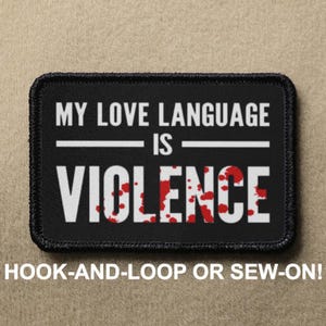 May include: Black patch with white text that reads "MY LOVE LANGUAGE IS VIOLENCE" with red splatter paint. The patch has black stitching around the edges and says "HOOK-AND-LOOP OR SEW-ON!" below.