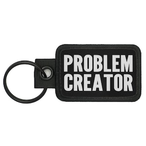 Könnte beinhalten: Schwarzer Schlüsselanhänger mit einem schwarzen rechteckigen Anhänger, auf dem "PROBLEM CREATOR" in weißen Buchstaben steht.