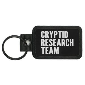 May include: Black key chain with a black rectangular tag that says "CRYPTID RESEARCH TEAM" in white text.