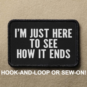 May include: Black patch with white text that reads "I'm just here to see how it ends". The patch is for hook-and-loop or sew-on attachment.