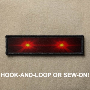 Puede incluir: Un parche rectangular negro y rojo con dos luces rojas brillantes. El parche tiene costuras negras alrededor de los bordes y el texto "HOOK-AND-LOOP OR SEW-ON!" está impreso debajo del parche.