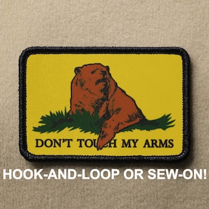 May include: A yellow patch with a black border features a cartoon brown bear sitting on a green patch of grass. The text "DON'T TOUCH MY ARMS" is printed in black below the bear.