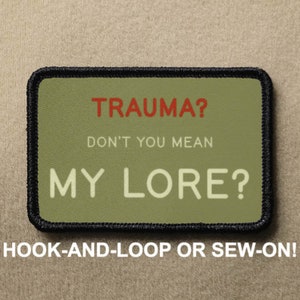 Op de afbeelding: Een groene en zwarte geborduurde patch met de tekst "TRAUMA? DON'T YOU MEAN MY LORE?" in witte en rode letters. De patch is ontworpen om met klittenband te worden bevestigd of aangehecht.