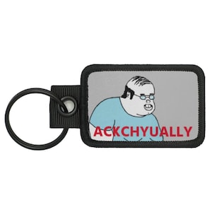 Könnte beinhalten: Schlüsselanhänger mit einem rechteckigen Aufnäher mit einer Cartoonfigur in einem blauen Hemd und einer Brille. Der Text "ACKCHYUALLY" ist rot aufgedruckt. Der Aufnäher hat einen schwarzen Rand und ist an einem schwarzen Schlüsselring befestigt.