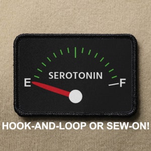 Puede incluir: Medidor blanco y negro con una aguja roja que apunta a "E" en una escala etiquetada como "SEROTONIN". El medidor está en un parche negro con costuras negras.