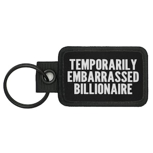 Puede incluir: Llavero negro con una etiqueta rectangular negra que dice "Temporarily Embarrassed Billionaire" en letras blancas.