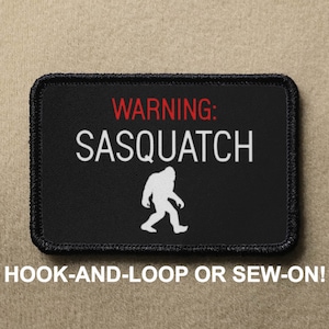 May include: Black patch with white text that reads "WARNING: SASQUATCH" and a white silhouette of Bigfoot walking. The patch has black stitching around the edges and says "HOOK-AND-LOOP OR SEW-ON!" in white text below the patch.