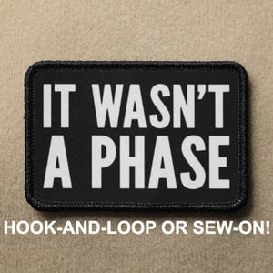Pode incluir: Etiqueta bordada preta e branca com o texto "IT WASN'T A PHASE" em letras brancas. A etiqueta tem uma borda preta e é projetada para ser fixada com velcro ou costurada.