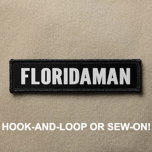 May include: Black patch with white text that reads "FLORIDAMAN". The patch has a hook-and-loop or sew-on backing.