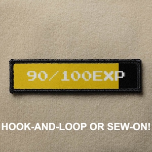 Könnte beinhalten: Gelber und schwarzer gestickter Aufnäher mit weißem Text, der "90/100EXP" lautet. Der Aufnäher ist zum Annähen oder Befestigen mit Klettverschluss konzipiert.