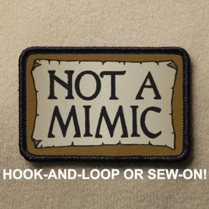May include: A brown and black patch with a white background and the text "NOT A MIMIC" in black lettering. The patch has a black border and is designed to be sewn or attached with hook-and-loop.
