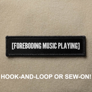 Pode incluir: Patch preto com texto branco que diz "[FOREBODING MUSIC PLAYING]" O patch tem um suporte de gancho e laço e pode ser costurado.