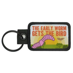 Può includere: Un portachiavi nero con un patch nero e bianco che dice "The early worm gets the bird". Il patch presenta un verme rosa e un uccello giallo.