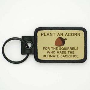 Può includere: Un portachiavi nero e marrone con un anello nero e una toppa marrone. La toppa ha il testo "PLANT AN ACORN FOR THE SQUIRRELS WHO MADE THE ULTIMATE SACRIFICE" e un'immagine di una ghianda marrone.