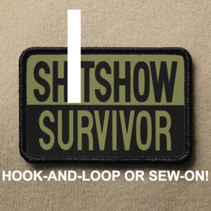 Könnte beinhalten: Schwarzer und grüner gestickter Aufnäher mit dem Text "SHITSHOW SURVIVOR" in weißen Buchstaben. Der Aufnäher hat einen schwarzen Rand und ist zum Anbringen mit Klettverschluss oder zum Annähen gedacht.