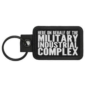 Puede incluir: Llavero negro con una etiqueta blanca que dice "Here on behalf of the Military Industrial Complex".