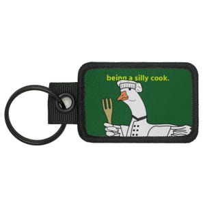 May include: A keychain with a green rectangular patch featuring a cartoon goose in a chef's uniform holding a fork. The text "being a silly cook" is above the goose. The keychain has a black ring and a black fabric loop.