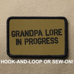 May include: A rectangular patch with the text "GRANDPA LORE IN PROGRESS" in black stencil font on a tan background. The patch has a black border and the text "HOOK-AND-LOOP OR SEW-ON!" below the main text.