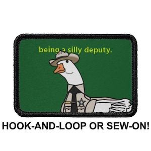 Puede incluir: Un parche bordado verde y negro con un ganso de dibujos animados que lleva un sombrero de sheriff y un uniforme. El texto "being a silly deputy." está por encima del ganso. El parche es para fijación de gancho y bucle o para coser.