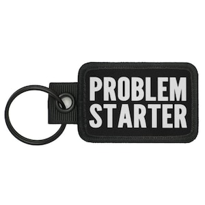 Könnte beinhalten: Schwarzer Schlüsselanhänger mit einem schwarzen rechteckigen Patch, auf dem "PROBLEM STARTER" in weißer Schrift steht.