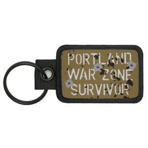May include: A rectangular keychain tag with the text "PORTLAND WAR ZONE SURVIVOR" in white. The tag is olive green with black accents and bullet hole graphics. A black key ring and strap are attached.