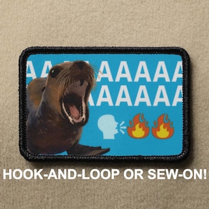 Op de afbeelding: Rechthoekige patch met een zwarte rand met een bruine zeeleeuw met open mond op een blauwe achtergrond. Witte tekst luidt "AAAAA" en "AAAAA". Daaronder staan spraak- en vuuremojis. De tekst "HOOK-AND-LOOP OR SEW-ON!" staat onderaan.