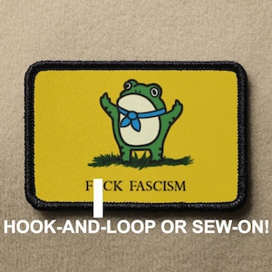 Peut inclure: Un écusson rectangulaire avec un fond jaune et une bordure noire. L'écusson représente une grenouille de dessin animé avec un nœud papillon bleu et le texte "F CK FASCISM". Le bas de l'écusson indique "HOOK-AND-LOOP OR SEW-ON!"