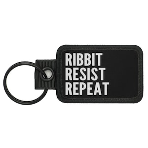 May include: Black rectangular keychain with the words "RIBBIT RESIST REPEAT" in white block letters. The keychain has a black metal ring and a black fabric loop.