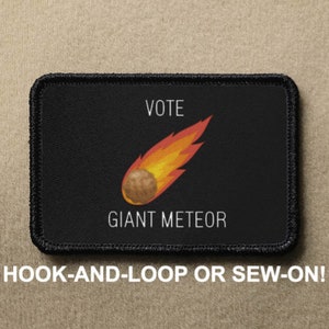 Könnte beinhalten: Schwarzer Patch mit weißem Text, der "VOTE GIANT METEOR" lautet, und einem Cartoon-Bild eines feurigen Meteors. Der Patch hat schwarze Nähte an den Rändern.