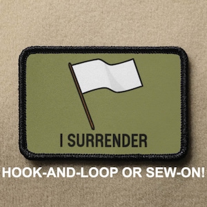 May include: A green and black embroidered patch with a white flag on a stick and the text "I SURRENDER". The patch is for hook-and-loop or sew-on attachment.