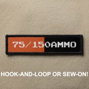 Könnte beinhalten: Ein schwarzer und orangefarbener Patch mit weißem Text, der "75/150 AMMO" lautet. Der Patch ist so konzipiert, dass er mit Klettverschluss befestigt oder angenäht werden kann.