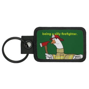 May include: A black key chain with a green and yellow patch featuring a cartoon goose wearing a firefighter's hat and holding an axe. The text on the patch reads "being a silly firefighter."