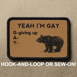 Op de afbeelding: Een bruine patch met zwarte stiksels heeft de tekst "YEAH I'M GAY" met het acroniem "G-giving up A- Y-" en een illustratie van een zwarte beer. De patch is gelabeld met "HOOK-AND-LOOP OR SEW-ON!"