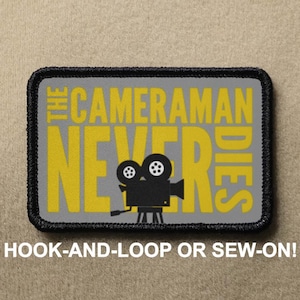 May include: A gray and black embroidered patch with yellow text that reads "The Cameraman Never Dies" and a black silhouette of a vintage movie camera. The patch has a black border and is designed to be sewn or attached with hook and loop.