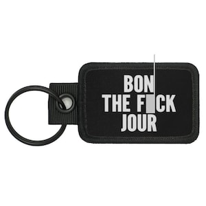 Puede incluir: Llavero negro con una etiqueta rectangular negra que dice "Bon the f*ck jour" en letras blancas.